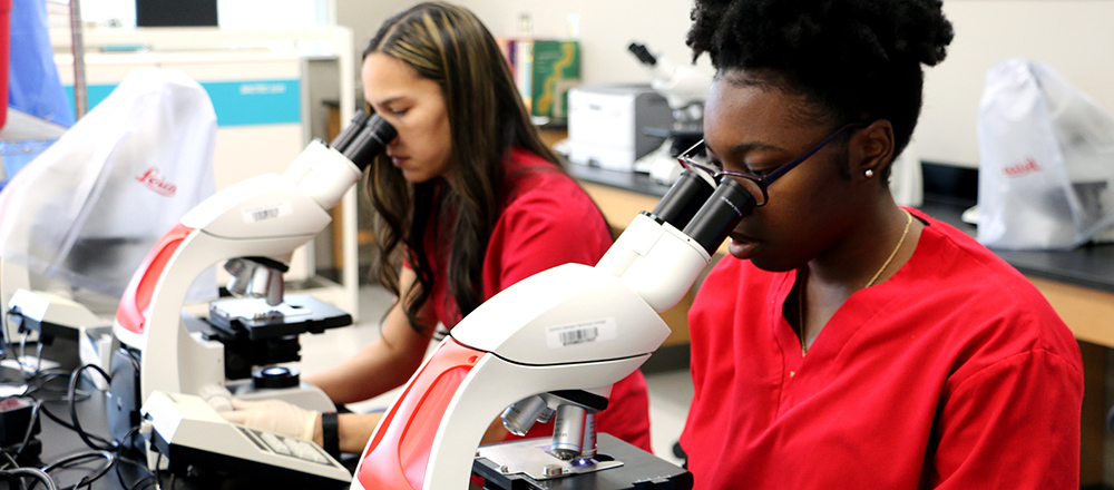 Two students in red scrubs, each looking through the lens of a microscope.