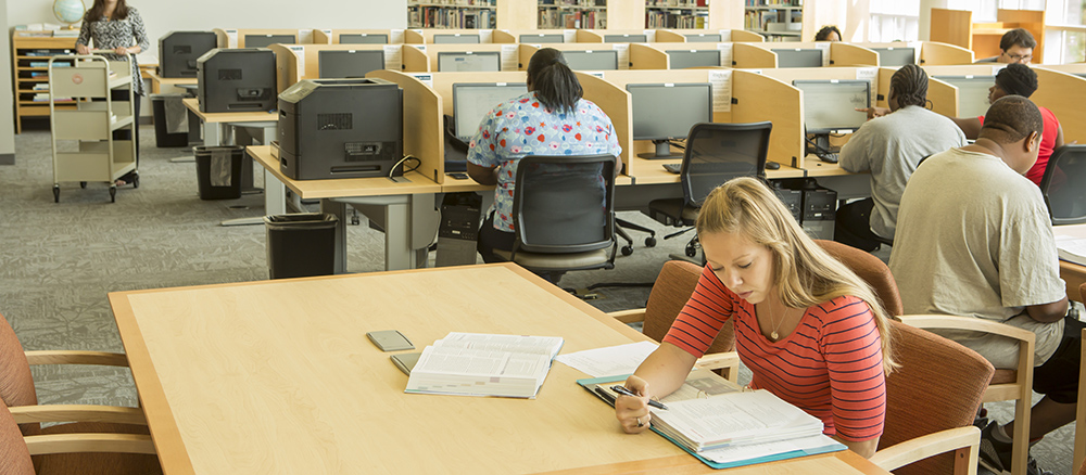Young woman sitting at desk in library reading a text book while several other students work on computers in the background in a library.
