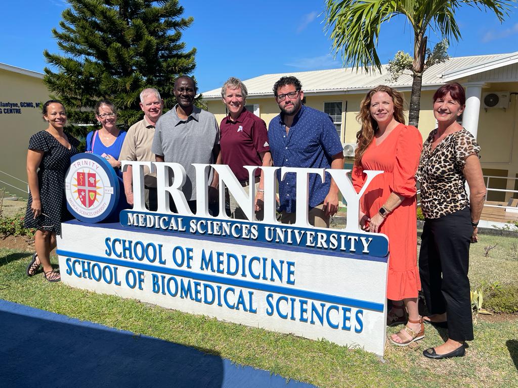 Leadership from CGTC and Trinity stand by Trinity School of Medicine sign outside its campus. 