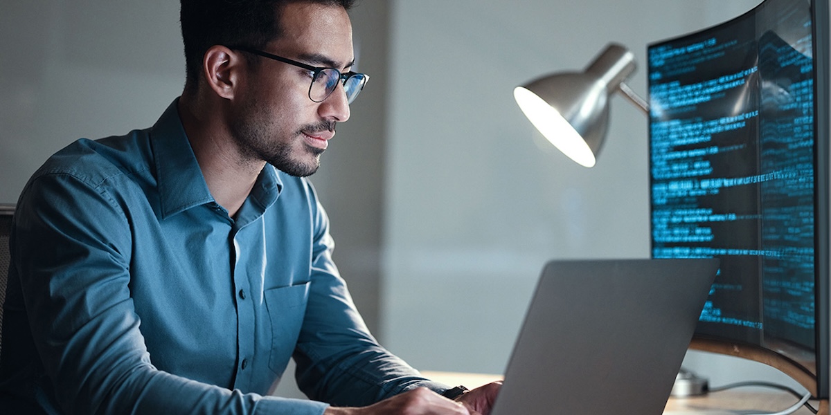 Man with glasses working on laptop at desk with an external monitor displaying code